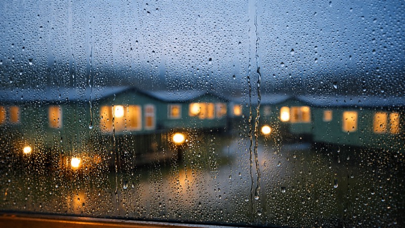 View through a rain-covered window looking out at static caravans with warm lights glowing at dusk