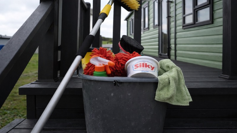 Cleaning supplies in a bucket on static caravan decking — mop, cloths, spray bottles and brushes ready for a maintenance session