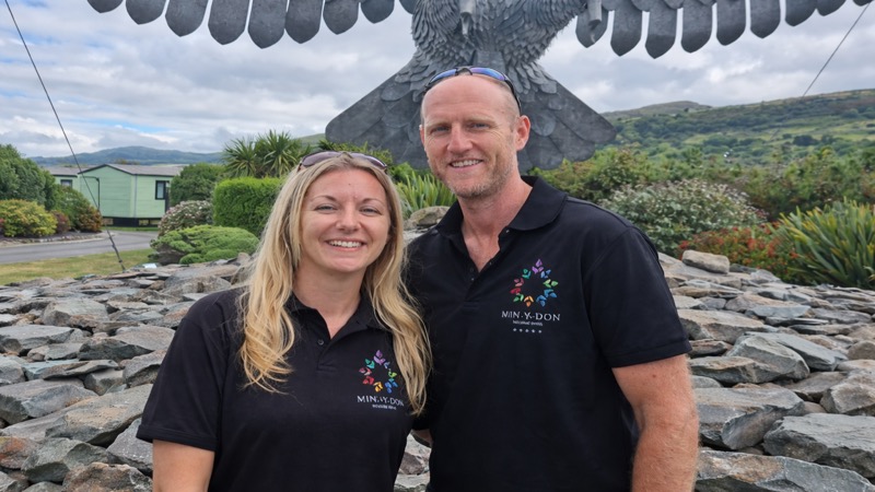 Park managers at Min-Y-Don Holiday Park, smiling in branded polo shirts with hills and holiday homes behind them